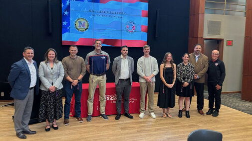 Group of people standing indoors in front of a screen displaying patriotic graphics at the MVSC honors ceremony.