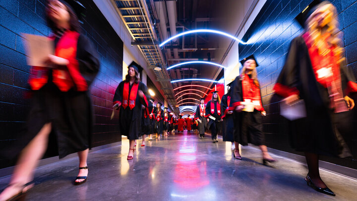 Graduates in caps and gowns walking down a hallway with colorful lights overhead.