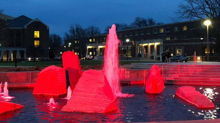 Large red-lit rocks and fountain sprays in a water feature at dusk with buildings in the background.