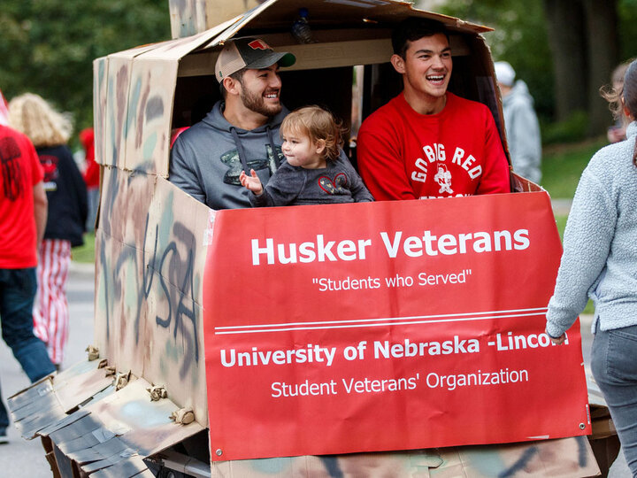 Husker Veterans parade float with people in camo-colored structure and red banner in outdoor setting.