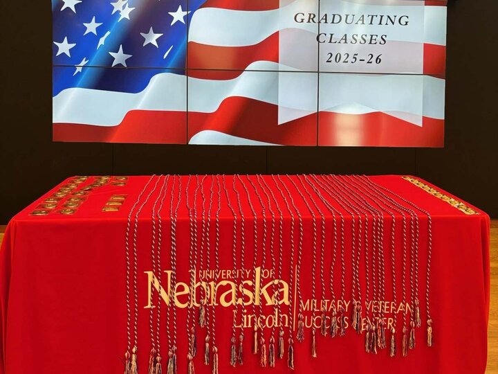 Red tablecloth with cords and medals, backdrop shows U.S. flag and text about graduating classes 2025-26.