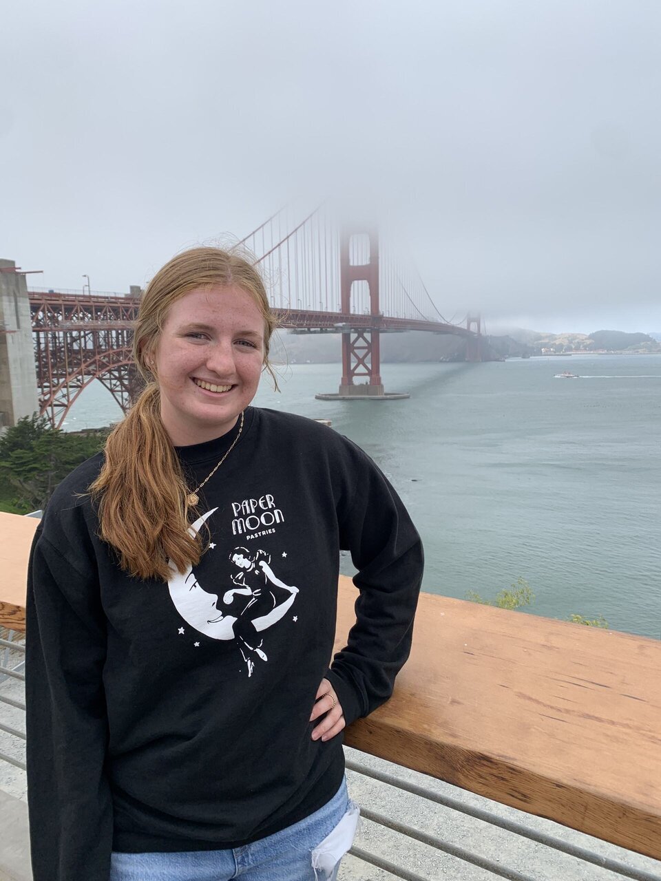 Person with a black sweatshirt stands near wooden railing, Golden Gate Bridge in foggy background.